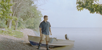 still from 'Ozigwan' by Cole Forrest picturing a man in rubber boots in front of a boat and a lake
